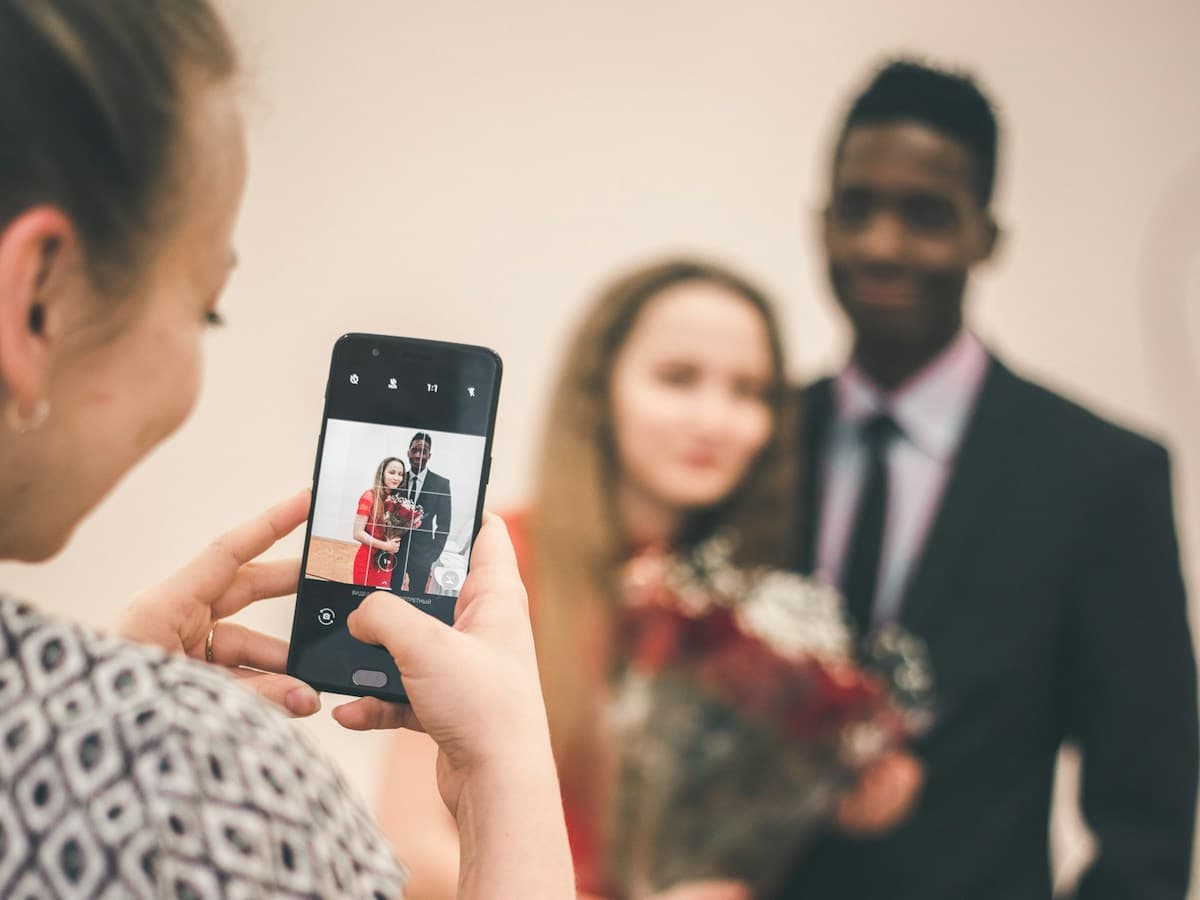 Convidada fotografa pelo celular uma jovem de vestido vermelho e um rapaz de terno